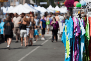 A street festival scene with a rack of colorful tie-dye shirts in the foreground. In the background